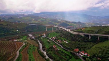 Concrete highway bridge stretching terraced hills in rural valley. Drone view asphalt road with vehicle crossing green landscape. Countryside farmland creating peaceful scenery under cloudy horizon.