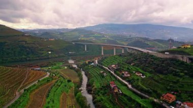 Scenic panorama rural nature under cloudy sky. Aerial view picturesque agricultural fields covering bright green slopes gloomy day. High bridge stretching over countryside hills with moving transport.