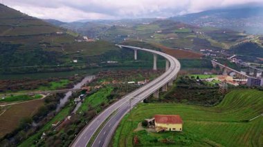 Drone view curved highway bridge rising on farmland in hilly valley. Concrete high road stretching on terraced slopes under cloudy sky. Transport crossing countryside landscape with village houses.