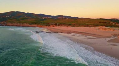 Aerial view scenic evening beach washed by turquoise sea water. Breathtaking marine landscape at golden sunset light. Foamy waves rolling on empty sand shoreline. Green hills raising on calm horizon.
