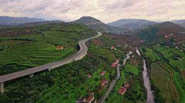 Stunning countryside greenery aerial view. Curve asphalt road running vivid slopes under cloudy sky. Small stream crossing lush green fields with village buildings. Picturesque gloomy rural landscape.