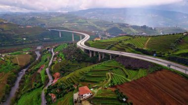 Elevated road curving hillside farmland with terraced slopes. Drone view concrete bridge over river village houses. Vehicles moving highway countryside over agricultural fields. Engineering structure.
