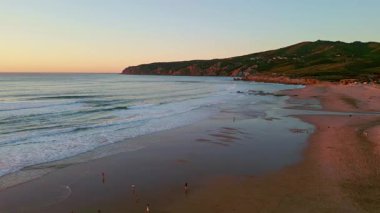Twilight beach landscape drone view. Calm ocean waves washing sandy wide shoreline in serene evening. Beautiful green mountain raising on horizon. Yellow skyline meeting deep sea surface at sunset.