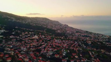 Aerial view evening Madeira settlement on ocean shore. Beautiful cozy streets with tiled roofs houses in twilight. Red buildings rooftops forming pattern on hillside. Architectural community style.