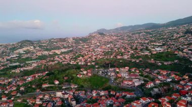 Dense Portuguese urban district surrounded lush greenery aerial view. Beautiful Madeira town under evening sky. Cozy streets stretching at hillside with tiled roof buildings. Picturesque panorama.