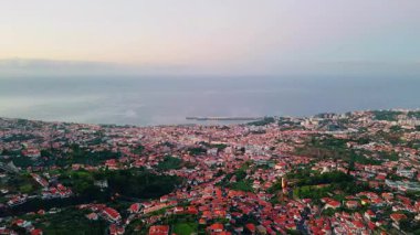 Aerial view picturesque coastal city at evening. Red rooftops stretching at urban slope. Cozy streets running through dense residential zone. Town architecture in beautiful ocean coast landscape.