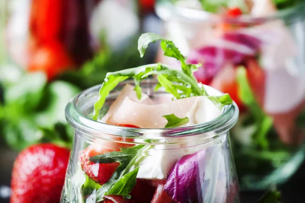 Siyah masada, seçici odak yaz salatası roka, yumuşak peynir, kırmızı çilek ve prosciutto cam kavanozlar