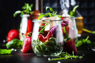 Siyah masada, seçici odak yaz salatası roka, yumuşak peynir, kırmızı çilek ve prosciutto cam kavanozlar