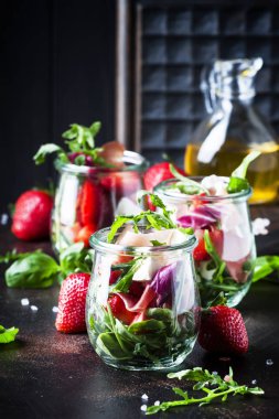 Siyah masada, seçici odak yaz salatası roka, yumuşak peynir, kırmızı çilek ve prosciutto cam kavanozlar