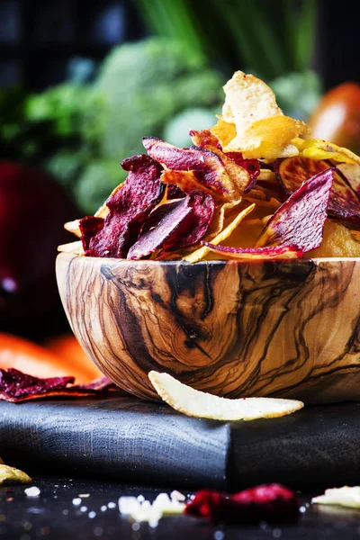 Vegan snacks, multicolored vegetable chips in  wooden bowl, background from  set of fresh farmer vegetables, still life, selective focus