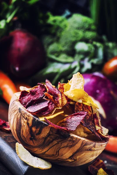 Vegan snacks, multicolored vegetable chips in  wooden bowl, background from  set of fresh farmer vegetables, still life, selective focus