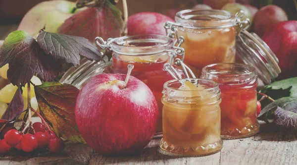 Homemade apple jam in jars in the autumn background with ripe apples and red leaves on the old wooden table