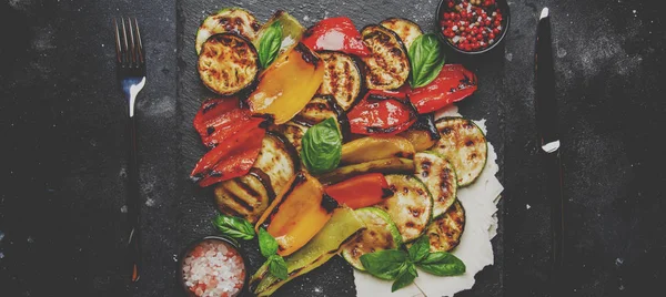 Grilled multicolored vegetables, aubergines, zucchini, pepper with green basil on serving stone board on gray background, top view