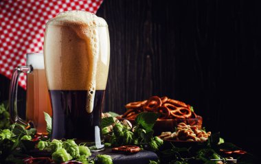 Dark German beer is poured into a glass, fresh green hops and bowls with salty snacks and nuts, autumn beer festival concept, dark background, selective focus clipart