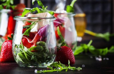 Baharatlı roka, keçi peyniri, çilek ve jambonlu taze salata veya modern cam kavanozlarda siyah masada seçici bir odak noktası.