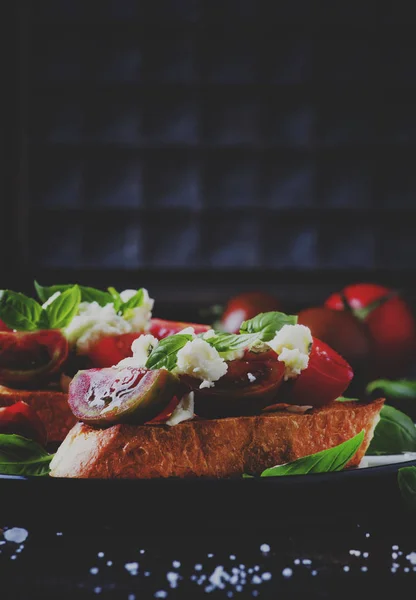 Crispy bruschetta with red and brown cherry tomatoes, feta cheese and green basil on the plate, selective focus