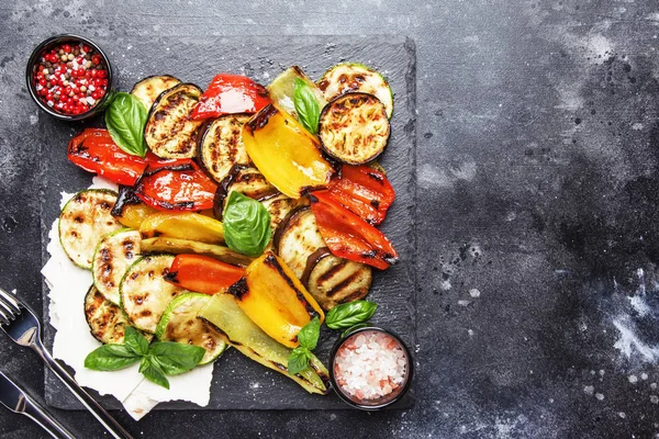 Grilled multicolored vegetables, aubergines, zucchini, pepper with green basil on serving stone board on gray background, top view
