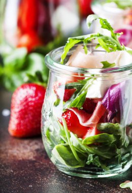 Baharatlı roka, keçi peyniri, çilek ve jambonlu taze salata veya modern cam kavanozlarda siyah masada seçici bir odak noktası.