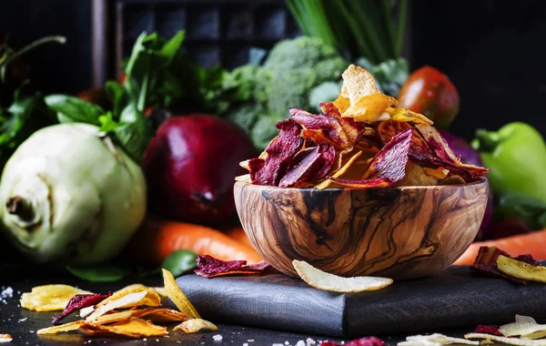 Vegan snacks, multicolored vegetable chips in wooden bowl and set of fresh farmer vegetables, rustic still life, selective focus
