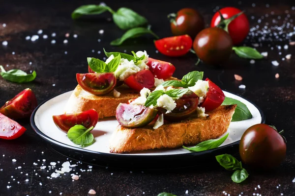 Crispy bruschetta with red and brown cherry tomatoes, feta cheese and green basil on the plate, selective focus