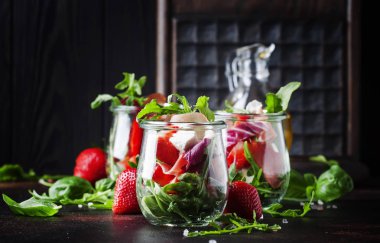 Baharatlı roka, keçi peyniri, çilek ve jambonlu taze salata veya modern cam kavanozlarda siyah masada seçici bir odak noktası.