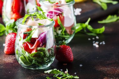Baharatlı roka, keçi peyniri, çilek ve jambonlu taze salata veya modern cam kavanozlarda siyah masada seçici bir odak noktası.