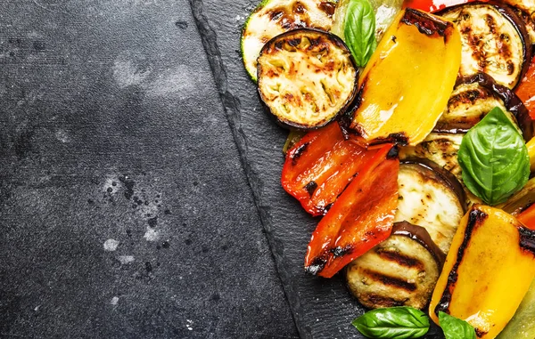 Grilled multicolored vegetables, aubergines, zucchini, pepper with green basil on serving stone board on gray background, top view