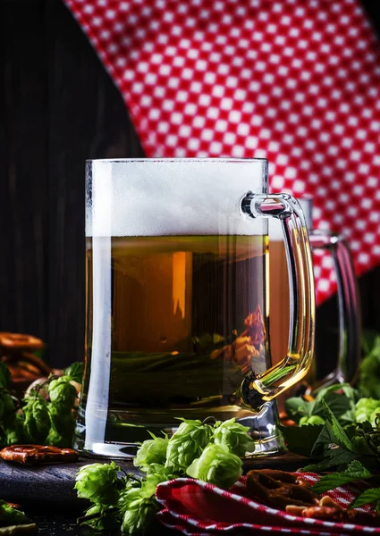 Light german beer poured into big glass, fresh green hops and bowls with salty snacks and nuts, autumn beer festival concept, dark background, selective focus