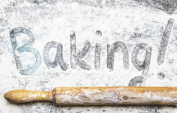 Food or baking background, white flour sprinkled on dark table, rustic wooden rolling pin, with inscription baking, top view