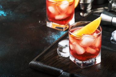 Summer alcoholic cocktail Negroni with dry gin, red vermouth and red bitter, orange slice and ice cubes. Brown bar counter background, bar tools, place for text, selective focus clipart