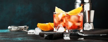 Summer alcoholic cocktail Negroni with dry gin, red vermouth and red bitter, orange slice and ice cubes. Brown bar counter background, bar tools, place for text, selective focus banner  clipart