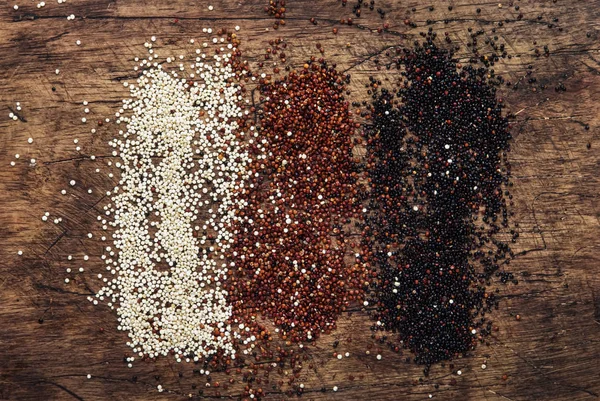 Black, white and red quinoa in spoons, raw quinoa groats assorted, wooden rustic kitchen table, top view