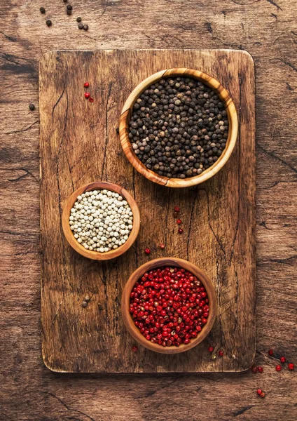 Black, white and pink rose peppers in bowls, assorted spices and spicy herbs on wooden rustic kitchen table, copy space, top view