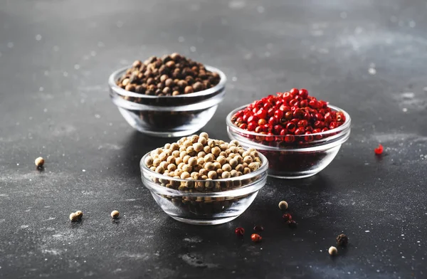 Black, white and pink rose peppers in bowls, assorted spices on gray kitchen table, selective focus