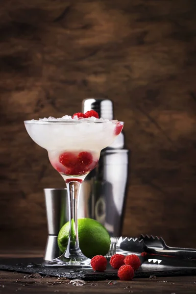 Raspberry daiquiri, alcoholic cocktail with white rum, lime juice, raspberries and crushed ice in tall glass, on wooden bar counter  with steel bar tools
