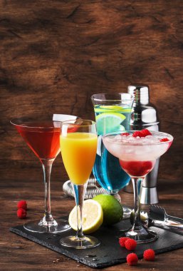Renkli yaz kokteylleri. Soğuk alkollü içecekler ve içecekler: mimoza, kozmopolit, ahududu margarita ve bar araçları ile ahşap masa arka plan üzerinde mavi hawaii