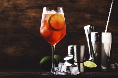 Kırmızı acı tonik, limon ve buzlu Campari tonik kokteyli. Eski ahşap masa arkaplanı, bar araçları, seçici odaklanma. Tüm isimler kokteyllere yöneliktir, markalara değil.
