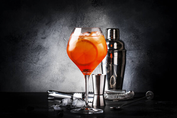 Aperol spritz cocktail in big wine glass with orange and ice, summer alcoholic cold drink, dark bar counter with steel bar tools, copy space
