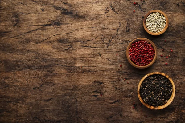 Black, white and pink rose peppers in bowls, assorted spices and spicy herbs on wooden rustic kitchen table, copy space, top view