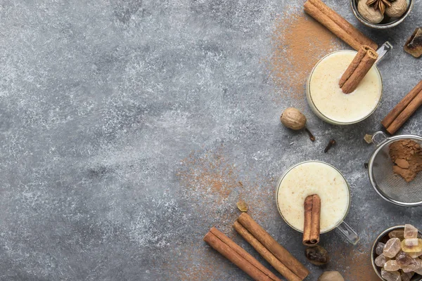 Eggnog hot winter or autumn drink with milk, eggs and dark rum, sprinkled with cinnamon and nutmeg in cups on gray background, copy space, top view