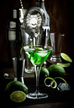 Bardakta yeşil martini alkollü kokteyl, sek cin, vermut, limon ve buz, çelik bar aletleri, koyu arka plan.