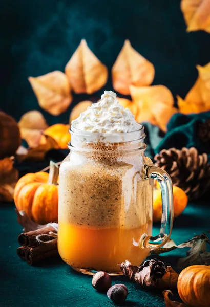 Pumpkin spiced latte or coffee in glass jar on blue table. Autumn or winter hot drink in festive natural table setting with orange leaves, spices, small pumkins, pine cones