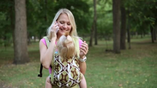 Una mujer lleva a un niño en un cabestrillo y habla por teléfono . — Vídeo de stock