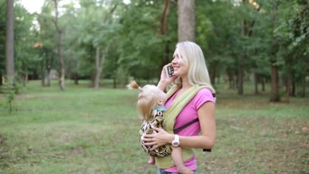 Uma mulher com um bebê em uma funda e falando ao telefone . — Vídeo de Stock
