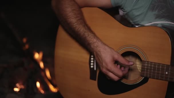 En man spelar en gitarr av branden på natten. — Stockvideo