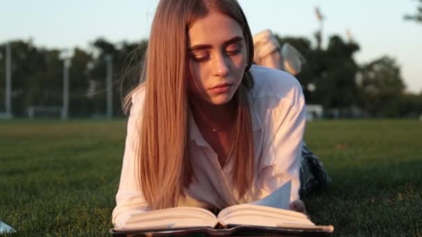 Uma bela jovem está lendo um livro no parque. Um estudante e uma estudante . — Vídeo de Stock