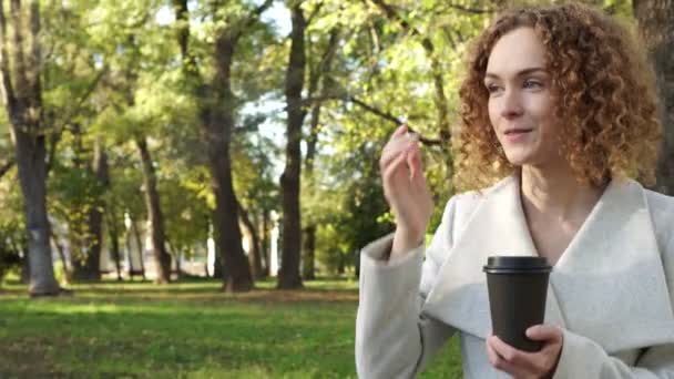 Mujer joven con el pelo rizado está tomando café en el parque de otoño . — Vídeo de stock