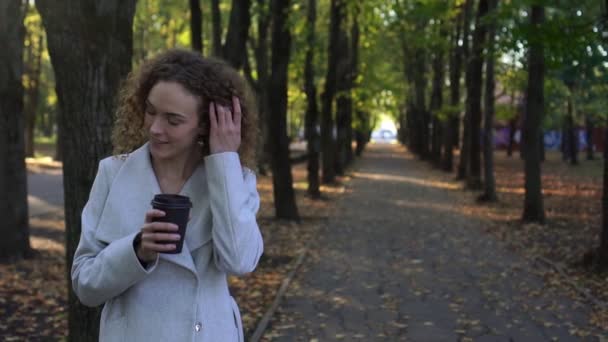 Krásná mladá žena s kudrnatými vlasy pije kávu v podzimní park. — Stock video