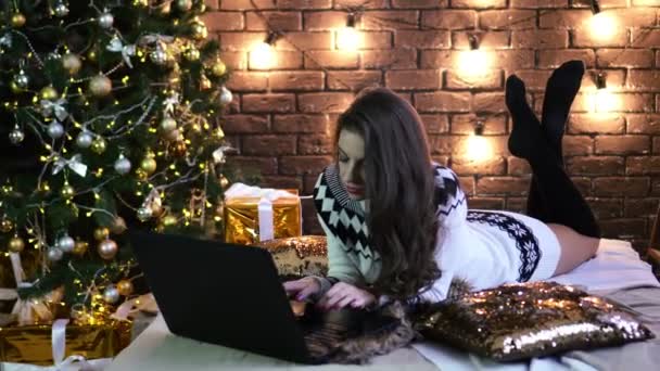 En ung kvinna med en laptop som ligger på sängen på bakgrunden av juldekorationer. — Stockvideo