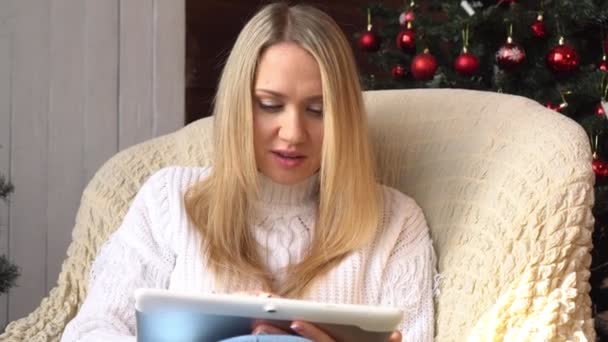 Uma mulher usa um computador tablet no fundo de uma árvore de Natal . — Vídeo de Stock